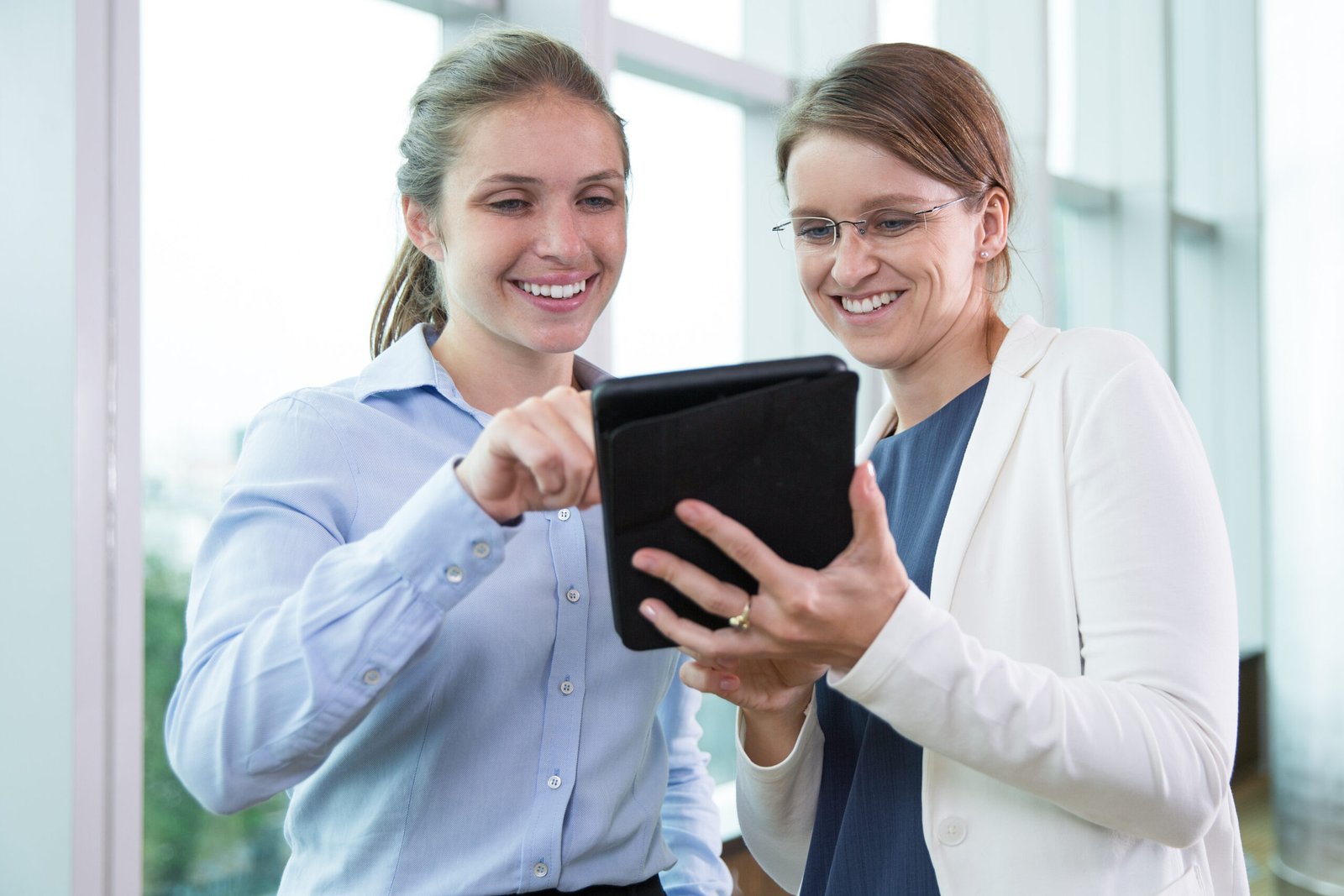 Portrait of cheerful female colleagues standing together, networking on touchpad and laughing in office